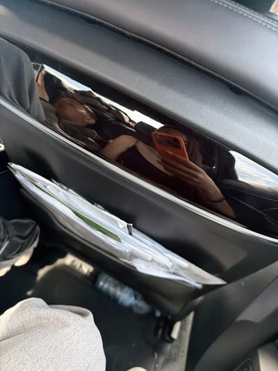 The image shows an interior car setting with a partial reflection of a person holding an orange phone, presumably taking the photo. Documents in envelopes are visible, stacked in the door pocket. Parts of a sneaker, a water bottle, and light gray clothing are also present. The location is not evident from the image.