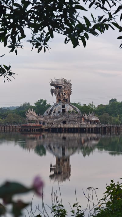 A full shot captures the Ho Thuy Tien Water Park, an abandoned water park structure designed to resemble a dragon, located in Thủy Xuân Ward, Vietnam. The dragon structure stands on a lake, its reflection visible in the water. The foreground features blurred foliage, while the background shows trees and a cloudy sky.