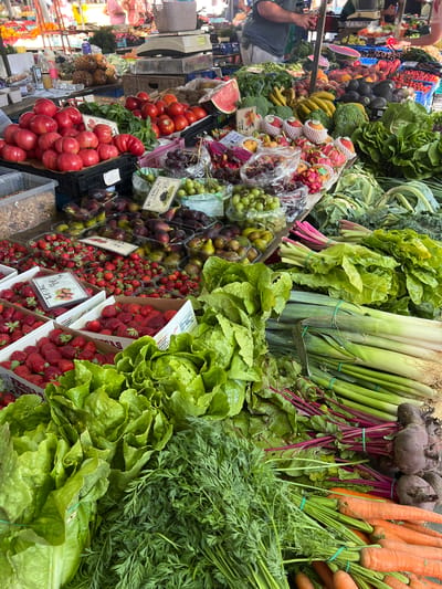 This is a high-angle, full shot of a vibrant produce stall at an outdoor market in Port de Pollença, Spain. The foreground is dominated by a large display of fresh produce. Bunches of bright orange carrots with green tops are stacked in the lower right, secured with green rubber bands. To their left are lush heads of green lettuce, also banded. Behind the lettuce, a pile of beets with deep purple stems and red roots is visible. Further back, a large bundle of leeks with white and green stalks, tied with green bands, is prominent. To the left of the leeks, leafy greens, possibly chard or kale, are arranged. In the midground, several boxes and crates overflow with various fruits and vegetables. Red tomatoes are piled high in a black crate, with a few bunches of basil interspersed. Nearby, containers of fresh strawberries are abundant, in various stages of arrangement. Green grapes are also visible in clear plastic containers. In the background, other market stalls are partially visible, with various fruits and vegetables on display, including what appear to be pineapples and possibly melons. People are present in the background, some tending to the stalls, and others browsing, though their faces are not clearly discernible. The overall impression is one of abundance and freshness at a bustling local market.