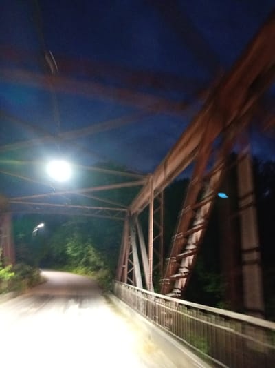 The image shows a road traversing a bridge at night. The bridge is constructed of metal beams, painted reddish-brown, and exhibits signs of rust. A bright light fixture, likely a streetlamp, illuminates the road. The road curves to the left as it enters the bridge structure. Dense foliage lines the sides of the road leading up to and surrounding the bridge. The sky above is dark blue, indicating nighttime. The overall scene suggests a rural or semi-rural location, possibly in Bgardvany, Abkhazia, given the prompt's context. The motion blur in the image indicates that the photograph was taken while the camera was moving.
