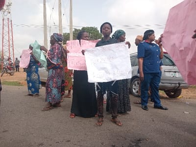The image shows a protest taking place in Ungwan Tanko, Nigeria, at coordinates 10.422 latitude and 7.49 longitude. There are eight individuals in the image. A group of women are holding signs in front of them, protesting against the government. They are standing in front of a silver car. A police officer stands to the right side of the image. Two other people, seemingly not involved in the protest, stand behind the women. One of the protesters is holding a sign that says "What Do We Get From This Government For Our Money?" in handwritten black ink. The ground is paved and the street is empty. The sky is cloudy and the sun is obscured. The image appears to be taken during the daytime.