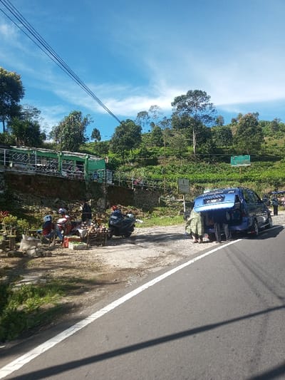 The image displays a roadside scene in Ka Thaung (Upper), Myanmar, under a clear blue sky with wispy white clouds. An asphalt road, marked by a solid white line on its right shoulder, extends into the mid-ground.
On the left side of the road, several individuals are positioned around makeshift roadside stalls on a dirt patch. Two people are discernible near the stalls: one wears a light-colored helmet and a red top, and another wears a dark top. The stalls are constructed from wooden tables and display various items, including what appears to be fresh produce in crates and sacks. Two motorcycles are parked adjacent to these stalls; one dark-colored motorcycle faces away from the viewer. Behind the stalls, a multi-level structure with green railings and a green roof is integrated into the hillside, with several people visible on its upper level. The hillside itself is densely covered with green vegetation, including numerous trees and what resembles a cultivated tea plantation.
On the right side of the road, a blue minivan with an open rear hatch is parked on the dirt shoulder. Two individuals are observed bending over, actively interacting with the contents of the minivan's rear compartment; one person wears a patterned garment, the other dark clothing. Further down the road on the right, additional vehicles and indistinct figures are present. Two signs are mounted on the hillside behind the minivan: one large green sign and a smaller, lighter-colored sign. Multiple black power lines are strung across the upper left quadrant of the sky. The scene is illuminated by bright daylight, casting shadows indicative of a high sun angle.
