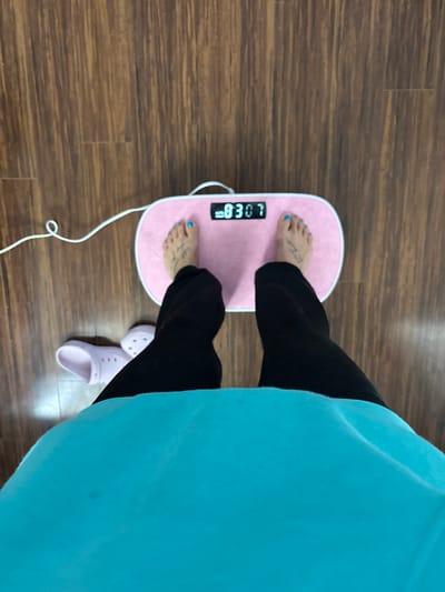 This overhead shot captures a person standing barefoot on a pink, oval-shaped foot massager, positioned on a dark brown wooden floor. The person is wearing black leggings or pants and a loose-fitting, light blue or turquoise top, which drapes down on the right side of the frame. Both of the person's feet are placed on the textured surface of the massager. The left foot is clearly visible, adorned with bright blue painted toenails and a small, partially legible tattoo on its top. The massager's digital display shows the numbers "83:07". A white power cord extends from the massager towards the bottom of the frame. Beside the person's right foot, on the wooden floor, rests a pair of light purple Crocs-style slip-on shoes. The setting appears to be an indoor space, possibly a home or wellness area, located in Seal Beach, United States of America. The activity suggests a moment of relaxation, self-care, or a fitness routine involving the foot massager.