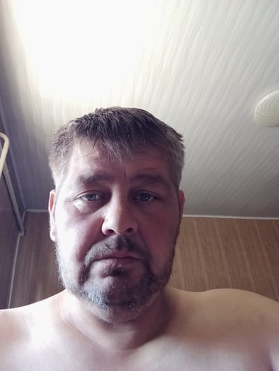 This is a close-up, selfie-style image of a middle-aged man with light-colored skin, short, dark brown hair with some graying, and a salt-and-pepper beard and mustache. He has blue eyes and is looking directly at the camera. The man is shirtless, and his shoulders are visible. The background consists of a wood-paneled wall and a white, textured ceiling with a light fixture visible on the right side. There is also a vertical, cream-colored pipe or railing on the left side of the image. The lighting suggests an indoor environment, possibly a bathroom or a room with wood paneling. The image quality is moderate, with some graininess. The scene does not contain any readily identifiable landmarks or context to suggest its specific location within the city of Esik, Kazakhstan.