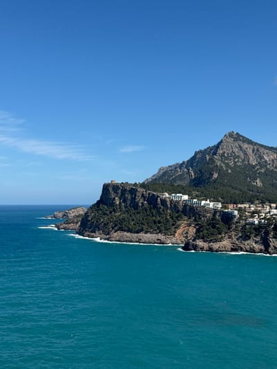 The image displays a coastal panorama near Port de Sóller, within the municipality of Sóller, Mallorca, Spain. The foreground consists of calm, turquoise-blue Mediterranean Sea with minimal surface agitation, featuring small white waves breaking against a rugged, dark rock shoreline. A prominent, tree-covered cliff rises steeply from the sea, topped by a circular, light-colored stone watchtower, identified as Torre Picada, situated on the left half of the upper cliff edge. To the right of the tower, a cluster of white and light-colored buildings, indicative of residential or resort development, are terraced into the vegetated slopes of the cliff face. Behind the coastal structures, a large, rugged mountain, part of the Serra de Tramuntana range, ascends into the background, covered in dense dark green vegetation on its lower slopes and exposing rocky outcrops near its peak. The sky overhead is clear and bright blue with a few wispy white clouds near the horizon on the left. No individuals, vehicles, or overt actions are visible in the scene.