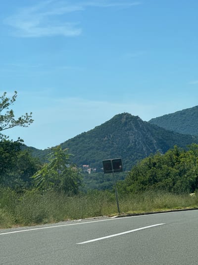 The image depicts a scenic view of a mountainous landscape near Golubovići, Montenegro. A paved road with a white lane marker runs diagonally across the lower part of the frame, suggesting a roadside perspective. To the left and right of the road, lush green vegetation and trees create a dense natural border.
In the middle ground, a prominent, tree-covered mountain dominates the scene. Its rocky peak is visible, and a small structure, possibly a tower or monument, can be seen at its summit. The slopes of this mountain are densely forested with various shades of green foliage. Further in the background, rolling hills covered in similar vegetation recede into the distance, culminating in a range of larger mountains. A small cluster of buildings, identifiable by their red rooftops, is nestled within the valley on the slope of one of the mountains, hinting at a settlement or village.
A road sign, featuring a dark rectangular panel with a red symbol and a white arrow, is positioned on a metal pole in the foreground, to the left of the central mountain. The sky above is a clear, bright blue with a few wispy clouds scattered across it, indicating a sunny day. The overall impression is one of natural beauty and tranquility, with the road and sign suggesting human presence within a natural environment.