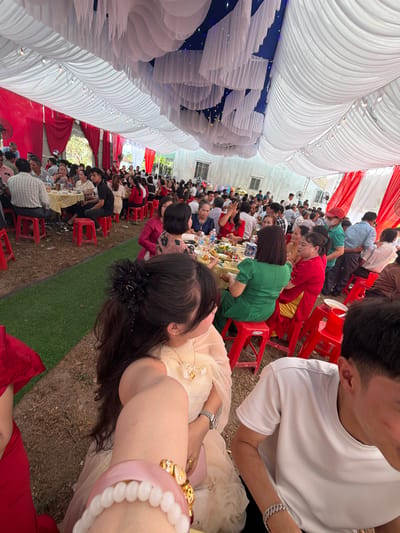 A high-angle view of a seated gathering of people under a tent-like structure in Tay Son Commune, Vietnam. The tent has white fabric draping and red and white accents, with a blue-lit section. Many people are seated at tables with food, sitting on red plastic chairs. The ground is a mix of grass and bare earth. Some people are interacting, while others are eating or looking at the camera. A person in a white shirt is visible in the upper right, and a person wearing several bracelets is prominent in the foreground.