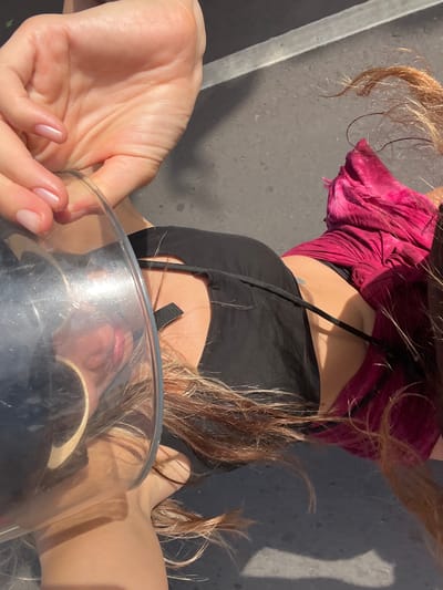 In Denuwala, Sri Lanka, a person's hand with painted fingernails holds a clear, curved object, possibly a glass or plastic bowl. The person is wearing a black garment, and a bright pink garment is visible on their right side. Their hair, a shade of brown with lighter streaks, is spread around them. The background appears to be a paved surface, possibly a street or sidewalk, with a faint linear marking. The lighting suggests it is daytime.