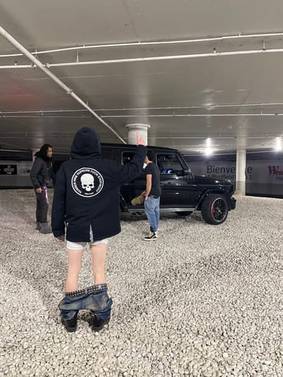 In a parking garage, a black G-Wagon is visible. Two individuals are near the car. One is adjusting something on it, while the other is reaching towards the ceiling. A person in a black hoodie faces away from the camera, pants around their ankles. Another person is standing further back in the frame. The floor is covered in small, light-colored rocks. Signage and parking garage infrastructure are visible.