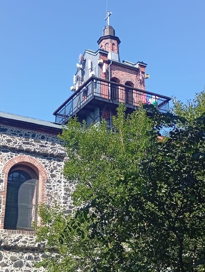 The image shows a close-up of the tower of a stone and brick building, likely a church or historical structure, against a clear blue sky. The tower is topped with a cross. Several modern antennas and communication equipment are mounted on the side of the tower and a balcony. A black metal railing surrounds a balcony on the tower. In the foreground, lush green trees partially obscure the view of the lower part of the building. The building has a large arched window with a metal grate. The architecture suggests a historical context, possibly within the city of Sobótka, Poland, known for its notable structures.