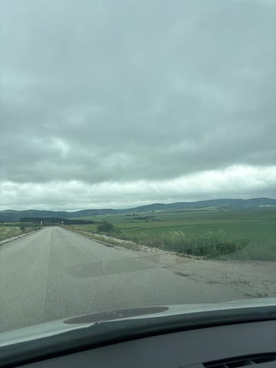A wide, paved road stretches forward under a heavily overcast, grey sky. The road is bordered on both sides by grassy verges and beyond that, expansive green fields that extend to a line of rolling, tree-covered hills in the distance. The overall scene suggests a rural or semi-rural landscape in Slivnitsa, Bulgaria, during a cloudy day. There are no people or vehicles visible on the road or in the fields.