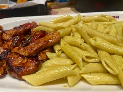 The image shows a close-up of a white plate containing two distinct dishes. On the left side of the plate are pieces of glazed chicken, appearing to be glazed with a dark, savory sauce that has a glossy finish. Some pieces show charring, suggesting they may have been grilled or pan-fried. Interspersed with the chicken are some red and green pieces, possibly bell peppers or other vegetables, coated in the same glaze.
On the right side of the plate is a generous portion of penne pasta. The pasta is cooked al dente and appears to be lightly dressed, possibly with a creamy sauce or olive oil and herbs, as evidenced by small green specks throughout. The penne pasta is cut diagonally at both ends and has ridges running along its surface.
The setting appears to be indoors, likely a restaurant or a home dining area. In the background, out of focus, there are hints of other items on a table, including what looks like a black container with a red logo, and some blurred text on a purple surface, possibly part of a menu or packaging. The lighting suggests it could be daytime, or an indoor environment with good lighting. There are no people visible in the frame. The focus is entirely on the food presented on the plate. The image is a still shot, capturing a moment of food presentation. The provided location context of Bağcılar, Turkey, does not offer any direct visual cues in the image itself.