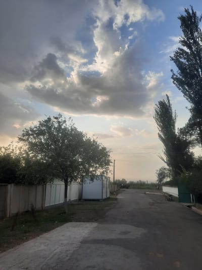 The image depicts an outdoor scene on a paved road in Voseaobad, Tajikistan. The sky is filled with dramatic, layered clouds, with patches of blue sky visible between them. On the left side of the road, a light-colored fence runs along the property line, with a small, bare-trunked tree in front of it. To the right of the tree are two modular white structures. Further down the road, tall, slender trees with dark foliage are prominent on the right side. The road appears to be an asphalt surface with some lighter patches, suggesting it might be wet in places. In the distance, there are more trees and vegetation, and mountains are faintly visible on the horizon. A utility pole with multiple wires is visible near the center-left of the image. The overall impression is that of a rural or semi-urban setting with a dramatic sky.