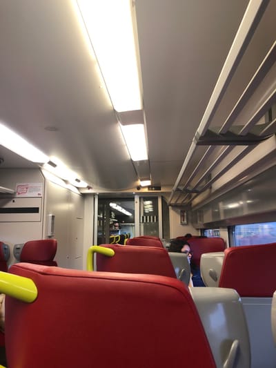 The scene depicts the interior of a modern passenger train carriage, purportedly located in the city of Hochfelden, France. The carriage is brightly illuminated by multiple elongated, recessed fluorescent or LED light fixtures on the light-colored, arched ceiling. On the right side, light-grey slatted luggage racks are visible above a row of windows, which show a blurred, indistinct exterior view.

The seating consists of numerous high-backed passenger seats upholstered in a vibrant red material, with grey or light-colored frames. Several seats, particularly in the foreground, feature bright yellow curved handles on the top edge of their backrests. The seats are arranged in rows, mostly facing forward.

One individual is visible seated on the right side of the carriage, approximately midway, with dark, curly hair and glasses, facing towards the window. A second, partially obscured head with dark hair is visible further down the aisle on the right. No direct interactions between individuals are discernible.

At the far end of the carriage, a doorway or passageway leads to another section of the train, with additional lighting visible within. Above this doorway, a white sign with upward and downward arrow icons is affixed. On the left wall, a white panel with a rectangular slot and a red and white sign (partially visible, resembling a "SNCF" logo with text "3117") are present. A red fire extinguisher or similar safety device is mounted on the wall panel on the right side, further down the carriage towards the window. The overall environment appears clean and functional.