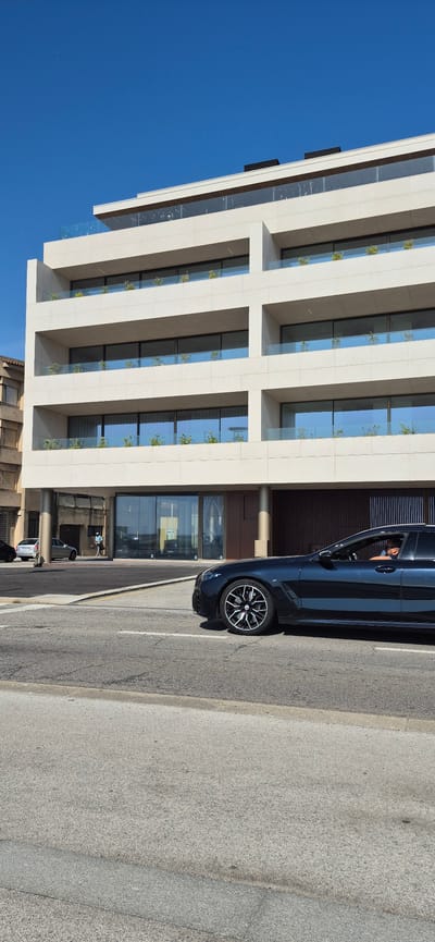 A navy blue car with a black interior and tinted windows is driving on a street in Vila do Conde, Portugal. The car is in the foreground, and its front passenger side is visible. The driver, a white male with dark hair and a beard, is wearing a black t-shirt and sunglasses. He is looking forward. The car has a black roof and a black trim around the windows. The wheels are black with silver spokes. Behind the car, there is a modern building with multiple levels. The building has a cream-colored facade with rows of balconies and large windows. Small plants are visible on the balconies. The sky is clear and blue. To the left of the building, another building with a darker facade and brown window frames is partially visible. In the background, there are two silver cars parked on the side of the street and a person walking. The street is paved with asphalt and has white lane markings.