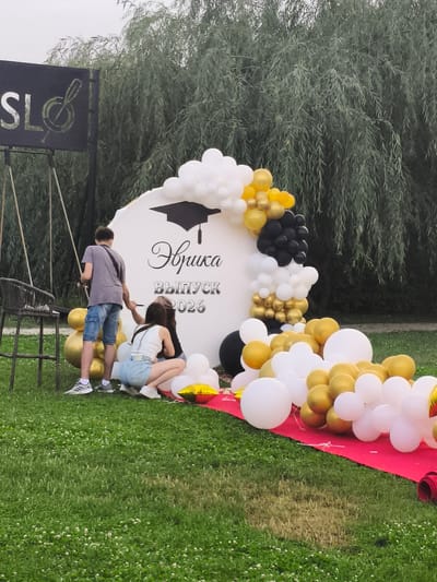 The image depicts a graduation-themed photo booth setup in an outdoor grassy area, possibly a park. A white, circular backdrop features a graduation cap illustration and Cyrillic text that reads "Эврика Выпуск 2026" (Eureka Graduation 2026). The backdrop is adorned with a balloon arch in white, gold, and black.

In the foreground, three young people are interacting with the setup. A young man in a grey t-shirt and denim shorts stands near the backdrop, holding a string. Two young women are kneeling and arranging balloons on the ground. One is wearing a white tank top and denim shorts, while the other has on a black top and jeans.

To the left of the backdrop, a dark, tall sign with a musical note symbol and the letters "SL" is visible. A dark chair with a woven back is placed nearby. The ground is covered in green grass with small white flowers. A red carpet is laid out in front of the backdrop, with more balloons scattered on it and the grass. In the background, tall trees with drooping branches, likely willows, create a lush green canopy.

The lighting suggests it is daytime, possibly overcast given the soft light and lack of strong shadows. The overall mood appears to be festive and celebratory, fitting for a graduation event. The location is identified as Komsomolskoe, Kyrgyzstan.