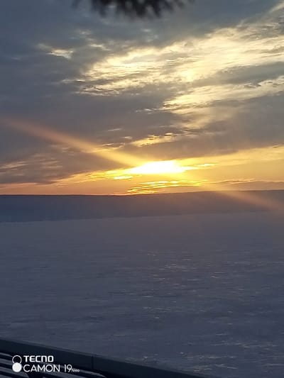 The image presents a winter landscape observed at either sunrise or sunset over a vast, frozen body of water near Klimovka, Russia, likely the Volga River or Kuybyshev Reservoir. The sky, occupying the upper portion of the frame, features scattered and striated clouds varying in color from dark grey on the left to intense yellow and orange in the center-right, where the sun, partially obscured, casts pronounced crepuscular rays. A narrow, dark horizontal band delineates the distant land or shore against the illuminated sky. The lower half of the photograph depicts a wide, flat expanse of bluish-grey, frozen surface, presumably ice or snow covering the water. A dark, horizontal railing or barrier is visible along the bottom edge, indicating the photograph was taken from an elevated viewpoint. Blurred, dark forms, possibly tree branches, are present at the top edge. No human figures, animals, or active subjects are discernible. The watermark "TECNO CAMON 19 Neo" is displayed in the lower-left corner.
