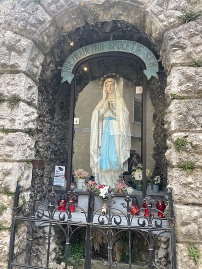 A ground-level, long-shot image depicts a small, enclosed shrine located in the city of Skofja Loka, Slovenia. The shrine is built into a recess within a wall constructed from rough-hewn stones. Inside the shrine, a statue of the Virgin Mary is visible, lying horizontally and covered in a white cloth with a blue sash. The statue is enclosed within a glass case, and the front of the shrine is secured with a decorative black metal gate. Several red candle holders are affixed to the gate, and flowers are placed inside the shrine. Above the glass case, a curved sign is mounted, bearing text in a foreign language, possibly Slovenian. The sign reads "BREZDEZNO SPOCETJE SEM." A small surveillance notice, potentially indicating video recording, is attached to the inside of the shrine.