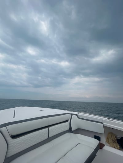 The image shows the back of a white boat with gray trim and a white, padded bench seat. The boat is moving forward on the water with the ocean in the background. The sky is overcast and gray. The location is on the Pacific Ocean near Playa Herradura, Costa Rica. The coordinates (latitude: 9.662, longitude: -84.707) place the location near the Puntarenas Province in Costa Rica. There are no people visible in the image. The boat is moving forward on the water, but the speed is not indicated. The conditions are overcast and gray. The sea is calm and blue. The boat's interior is clean and well-maintained. There are no signs of damage or wear. A tan towel is seen draped over the boat's rear corner.