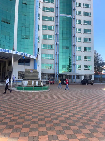 The image depicts an exterior view of the multi-story Alumni Plaza building of The Co-operative University of Kenya, located in Hospital ward, Kenya. The building features a light grey facade with extensive teal-tinted glass window panels and vertical blue decorative or structural elements. A prominent canopy over an entrance on the left reads "ALUMNI PLAZA" in dark blue lettering. Below the canopy, three flagpoles display flags, including one consistent with the Kenyan national flag.
In the foreground, a paved plaza composed of alternating reddish-brown and reddish-orange interlocking pavers extends towards the building. A circular planter with a green base and silver railing encloses a dark monument, which is inscribed with "THE CO-OPERATIVE UNIVERSITY OF KENYA" logo and text.
Six individuals are visible on the plaza. On the left, a male in a white shirt and dark trousers walks towards the left. In the midground, behind the monument, another male in a light-colored shirt is present near the building entrance. To the right, three individuals walk from right to left: a male in a red short-sleeved top and blue jeans, holding a mobile phone, flanked by two females wearing yellow tops, blue jeans, and white shoes. Another individual is partially obscured in the background.
Under a partial carport shelter on the right, a red car, a dark SUV, and a black pickup truck are parked. The scene is illuminated by bright daylight under a clear blue sky.