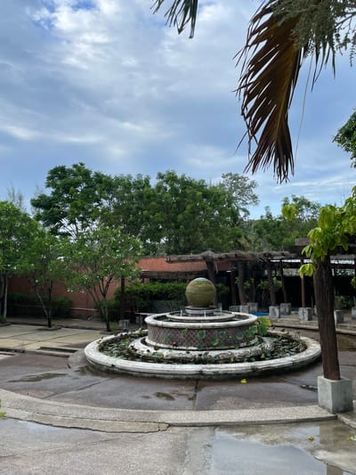 The scene is in Baan Wang Ta Kien, Thailand. A circular fountain with plants is positioned near a building structure with a wooden frame. Above, the sky is visible with wispy clouds, framed by foliage from trees and a palm leaf. The ground is paved, and portions appear wet, possibly due to recent rain.
