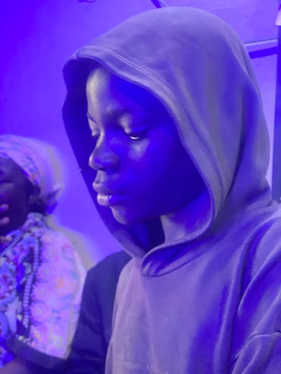 The image shows a close-up, vertically oriented shot of a person wearing a grey hooded sweatshirt. The person's face is visible in profile, looking downwards, with their face illuminated by a strong blue light that casts shadows and highlights. The hoodie is pulled up, obscuring most of their head. The background is predominantly a deep blue, with a hint of purple. In the lower right portion of the image, there are colorful patterned fabrics, suggesting the presence of other items or clothing. The overall lighting and color scheme create a dramatic and somewhat mysterious mood. There is no visible text in the image. The location context is Barakin, Nigeria.