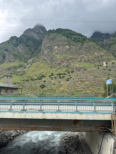 The image depicts a scene in Verkhnyaya Balkariya, Russia, featuring a rugged mountain landscape with steep, grassy slopes and rocky outcrops. A concrete structure, possibly a bridge or dam, spans a fast-flowing river. A blue metal railing runs along the edge of the structure, with a pathway visible behind it. The riverbank is lined with dark, rounded stones. Overhead, power lines crisscross the sky.

On the mountainside, several ancient-looking stone structures are visible, including what appears to be a tower and remnants of older buildings or fortifications. The slopes are a mix of green vegetation and bare rock. A flag with blue and white stripes is displayed near the top of the concrete structure.

The weather appears overcast, with a cloudy sky suggesting a cool, possibly damp day. The time of day is indeterminate due to the cloud cover. The overall impression is one of a remote, mountainous region with a sense of history evident in the ancient ruins. No people are visible in the image.