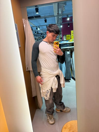 A man stands in a mirrored room, likely a clothing store fitting room. He is taking a selfie with a yellow smartphone. He is wearing a white and grey raglan shirt, dark grey jeans, and sneakers. He has a white sweatshirt tied around his waist. Garments hang on racks in the background. The room appears to be in a retail environment, possibly in Spreitenbach, Switzerland, given the presence of retail shelving, lighting, and mirrored walls typically found in clothing stores.