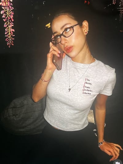 In a dimly lit indoor setting, possibly in the city of Korostyshiv, Ukraine, a young woman with dark hair pulled back is captured in a mid-shot. She is wearing clear-rimmed glasses and a light grey t-shirt with red text on it that reads "Vibe, Identity, Beauty, Aesthetics, Seduction". The woman is holding a mobile phone to her right ear with her right hand, and her left hand rests on her lap, adorned with a bracelet. Her left arm is also decorated with a pink beaded bracelet. To her left, a portion of a grey plaid jacket is visible, draped over something out of frame. The background is dark, with a blurred floral decoration on the left side and a few indistinct lights in the upper right quadrant, suggesting a possibly nighttime or dimly lit interior environment.