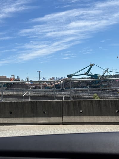 The image shows a coal loading facility in North Vancouver, Canada, under a blue sky with wispy white clouds. In the foreground, a concrete barrier partially obscures a highway or road. Behind the barrier, a metal railing separates the viewer from the industrial site. The site itself is dominated by large piles of coal, a massive teal-colored loading machine with conveyor belts, and various structures associated with industrial operations. In the far distance, a city skyline is visible, suggesting the proximity of a populated area. A bird is visible in flight in the upper right quadrant of the image. The scene appears to be taken during daylight hours.