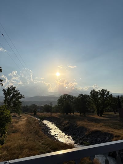 A scenic view of Mikrevo, Bulgaria, captures a sunlit waterway flanked by dry grass and trees under a vast, partly cloudy sky. The sun is positioned high in the sky, casting a warm glow on the scene and creating shimmering reflections on the water. Power lines are visible in the upper left corner, stretching across the frame. In the distance, rolling hills or mountains form a layered backdrop. The overall impression is one of serene natural beauty, possibly taken during a golden hour setting.