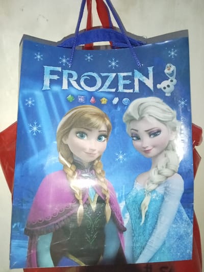 A blue, glossy paper gift bag featuring characters from Disney's "Frozen" is depicted in Inxuba Yethemba Nu, South Africa. The bag displays a blue background with white snowflakes and the word "FROZEN" in icy-textured white letters across the top center. Below the title are assorted colorful, gem-shaped graphics. The character Olaf, a white snowman, is visible in the upper right. The left side of the bag features an illustration of Anna, with light brown hair in two braids, wearing a magenta cloak over a dark dress. The right side shows Elsa, with blonde hair in a side braid, wearing a light blue gown. Both characters are depicted from the chest up, smiling slightly. The bag has two dark blue textile handles attached to its upper edge. A red object or surface partially frames the bag on the left and right sides. The background behind the bag is a light-colored, possibly off-white, wall, suggesting an indoor environment.
