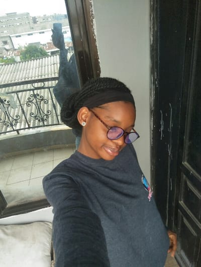 A young individual, appearing female, with dark skin and braided hair styled into an updo with a black headband, is centered in the image. She wears round, tortoise-shell framed glasses with blue-tinted lenses and a small, silver-colored stud earring in her visible left ear. She is dressed in a dark grey, long-sleeved t-shirt with a small, multi-colored emblem on the right chest. Her head is tilted slightly down and to her right, displaying a subtle smile. Her left arm, extended to hold a smartphone, is visible as a reflection in the glass panel to her upper left, indicating a selfie is being taken.

The scene is an interior setting with an urban background, specifically identified as Cotonou, Benin. To the individual's left, a glass door or window offers a view of the cityscape. Directly outside this glass is a black metal balcony railing with ornate scrollwork. Beyond the railing, numerous structures with corrugated metal roofs are visible, interspersed with trees. Further in the distance, multi-story concrete buildings rise under a bright, overcast sky. To the individual's right, a dark wooden door or door frame is partially visible. A light grey wall separates the door from the glass panel. A white pillow or cushion is partially visible in the lower left corner of the frame.