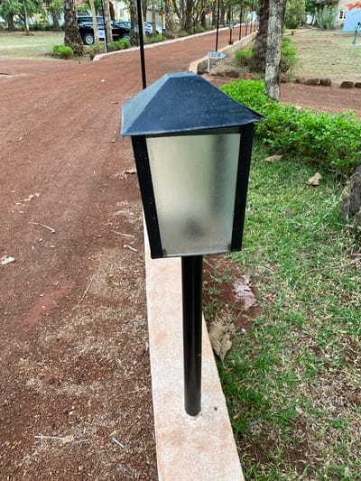 The image depicts an outdoor scene in Miango, Nigeria. A black, rectangular light fixture is the primary subject, mounted on a black pole and situated on a light-colored, possibly tiled, base. The ground is covered in reddish-brown soil interspersed with small pebbles and debris. Grass and some leafy plants are visible in the background, along with a line of trees in the distance, suggesting a park or garden setting. Cars are visible in the far background.