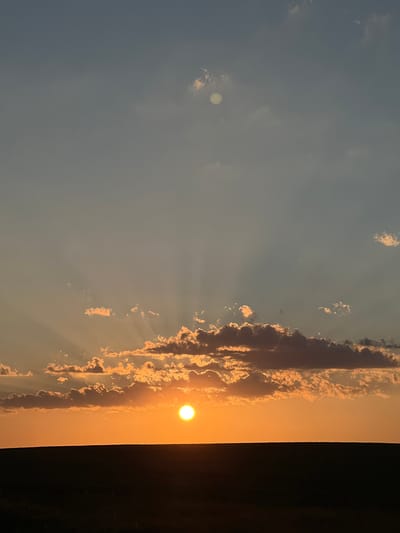 This is a vertical, eye-level shot of a sunset over Ilavche, Ukraine. The sun, a bright white orb, is visible in the upper right quadrant of the frame, casting an orange glow over the scene. Wispy clouds, illuminated by the sun, stretch across the sky from the right edge of the frame towards the left. Some clouds are darker, creating a contrast with the lighter ones. Sunbeams are subtly visible, radiating from behind the clouds. The sky transitions from a pale blue on the left to a warm orange on the right. The lower right edge of the image is dominated by a dark, indistinct area, possibly a tree line or building, that is in shadow. The overall mood is serene and tranquil, characteristic of a sunset. There is no visible text.