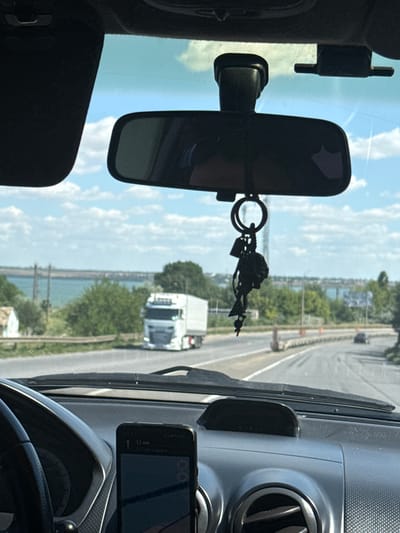 The image is a view from the interior of a vehicle, looking forward through the windshield and rearview mirror. The vehicle is on a road, with other vehicles visible ahead. In the distance, there is a body of water and a treeline. The sky is partly cloudy and blue. On the dashboard, a smartphone is mounted and displaying a navigation app. A keychain with a decorative pendant hangs from the rearview mirror. The scene appears to be a highway or main road, with some construction barriers present along the side of the road. The environment suggests a sunny day. The question of the location being in Usatove, Ukraine, cannot be verified from the image alone.