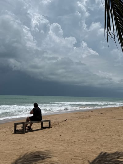 A person is sitting on a wooden bench on a sandy beach, facing the ocean. The sky is dramatic with dark, heavy clouds approaching from the left, contrasting with lighter, puffy clouds on the right. The ocean is a turquoise color with white waves breaking on the shore. A palm frond is visible in the upper right corner of the image. The beach is mostly empty, with the exception of the person on the bench and some scattered debris. The scene suggests an approaching storm. The location is the beach in Ban Khuk Khak, Thailand.