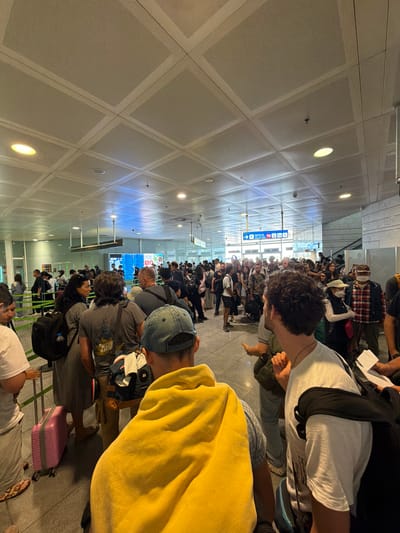 This image depicts a crowded airport security line at El Prat De Llobregat, Spain. A long queue of people, mostly young adults, are standing in line, some with luggage. The scene appears to be indoors, under a bright, artificial light, suggesting it could be daytime or evening. The ceiling is made of acoustic tiles, typical of an airport terminal. A blue and white sign with an arrow and an airplane icon is visible in the background, indicating the direction of the gate or a particular service. The atmosphere seems busy and possibly a little frustrating due to the wait. People are dressed casually, and some have backpacks or bags with them. A pink suitcase is visible in the foreground. The image is taken from a slightly elevated angle, looking down the line of people.