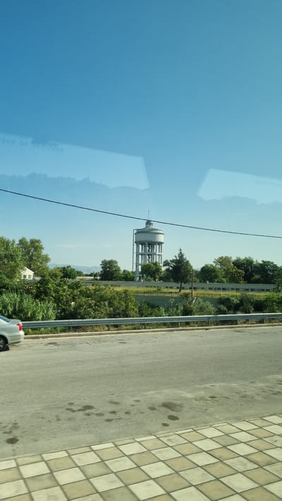 The image shows a water tower located in Larissa, Greece. The water tower is a tall, cylindrical structure supported by a network of beams and columns, with a domed roof. It is surrounded by lush green trees and vegetation. In the foreground, a road runs parallel to the scene, with a car partially visible on the left side. A metal guardrail separates the road from the grassy embankment. The sky is a clear, bright blue, with some reflections on what appears to be a window or glass surface in the upper part of the image, showing partial text that says "ISSUE DE SECOURS". The overall scene depicts a sunny day with a view of industrial infrastructure amidst natural greenery.