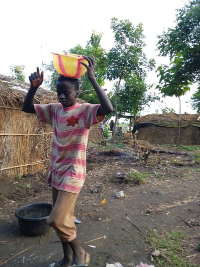 In Kajola, Nigeria, a young Black male with dark skin and short hair balances a yellow and pink striped bowl on his head. He is wearing a pink and beige horizontally striped t-shirt with a red emblem on the chest and light brown trousers. His arms are raised, with his hands positioned near the bowl, seemingly to help stabilize it. To his left, a rustic hut constructed from straw and bamboo is visible, with a rough, uneven ground covered in dirt, scattered straw, and debris in the foreground. Behind him, a large dark plastic basin filled with water sits on the ground. In the background, several trees with green foliage are scattered across the landscape, and more thatched structures, possibly huts or storage, are present. The sky is overcast and bright.