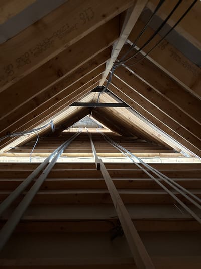 The image displays the unfinished interior of an attic or loft space within a building in Skipsea, United Kingdom. The view is directed upwards and towards the triangular apex of the roof structure. Numerous untreated wooden roof trusses and rafters form the primary A-frame framework. Stamped markings on several visible wooden beams include "James Jones," "1220-CPR-1036 UK," and "C16," indicating timber specifications and manufacturer. A dark metal beam or bracket is positioned horizontally near the apex, connecting opposing rafters. Electrical wiring, some bundled with zip ties, is routed along the wooden framework. Multiple white PVC pipes, likely conduits or plumbing lines, extend linearly across the lower half of the scene, parallel to exposed wooden floor joists. Natural light illuminates the distant end of the attic from an unseen opening. No individuals, tools, or personal effects are visible. The scene is static, depicting a construction or renovation phase.