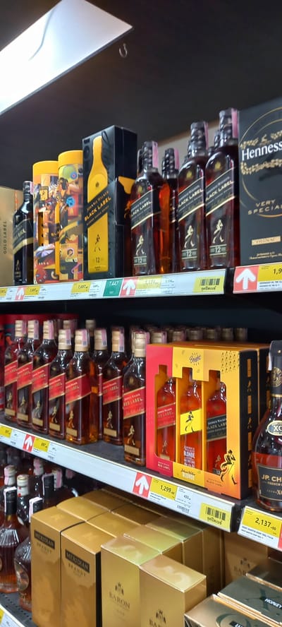 The image, rotated 90 degrees clockwise, captures a well-stocked aisle of alcoholic beverages in what appears to be a retail store, likely a supermarket or liquor store, in Moo 1, Thailand. The shelves are filled primarily with various brands and types of whiskey, brandy, and cognac.

On the left side of the frame, multiple bottles of Johnnie Walker Black Label whiskey are prominently displayed, stacked vertically. Price tags for these bottles show values of 1,174 and 1,697. Above these, a box for Hennessy Very Special Cognac is visible, with a partial price of 1,9__. Below the main stack of Black Label, there are special edition or gift-packaged Johnnie Walker Black Label products, including cylindrical packaging and boxes for Johnnie Walker Blonde, priced at 1,299.00, and another item at 433.

On the right side of the image, numerous bottles of Johnnie Walker Red Label are arranged on shelves, with a visible price of 697. Above them, boxed sets for Johnnie Walker Blonde and Johnnie Walker Red Label are stacked. Further up, a bottle of JP. Chenet French Brandy and a Remy Martin VSOP 70CL, priced at 2,137.00, are visible. Below the Red Label bottles, golden boxes for "Meridian Brandy" and "Baron Brandy" are neatly stacked, some with "HONEY" written on them.

The scene implies a retail environment focused on product display and sales. Many price tags feature an upward-pointing arrow and include Thai text, such as "จุดปิดเบย์" (Jut Pit Bay), which likely indicates a display point or special section, confirming the location in Thailand. The bottles and packaging are clean and well-organized, suggesting a commercial setting.