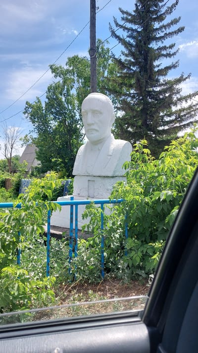 A bust of a man with a mustache and beard, made of white stone, stands on a pedestal in an outdoor setting. The bust is surrounded by lush green foliage, and a blue metal fence is visible in the foreground. Behind the bust, a wooden utility pole with wires runs vertically, and trees, including a tall evergreen, are present. In the background, a house with a grey roof can be seen. The sky is blue with scattered white clouds. The image appears to be taken from inside a vehicle, with the car's window frame visible along the bottom and right edge. This is a memorial bust located in the village of Topar, Kazakhstan.