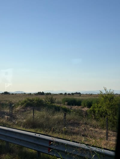 The image shows a wide, open landscape under a clear blue sky. In the foreground, a metal guardrail runs horizontally, partially obscuring a dry, grassy field. Several wooden posts, likely part of a fence, are visible within the field. The midground features a dense collection of green bushes and trees. In the distance, rolling hills are visible, with a hazy line of mountains on the horizon. The overall impression is of a rural or semi-rural setting, possibly a roadside view in the countryside. The image does not contain any people or specific identifiable structures within the city of Tsalapitsa, Bulgaria, as the setting is a general landscape view.