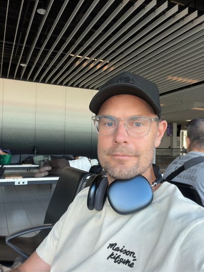 A man wearing a black baseball cap, clear-rimmed glasses, and a light-colored t-shirt with the text "Maison Kitsuné" embroidered on it, is taking a selfie. He has headphones draped around his neck. He appears to be in an airport waiting area, with rows of seating and other passengers visible in the background. One person is lying down on a bench, and another person's back is visible in the foreground to the right. The ceiling of the waiting area features a modern design with metal slats. This location is likely Schiphol Airport, situated in or near Badhoevedorp, Netherlands.