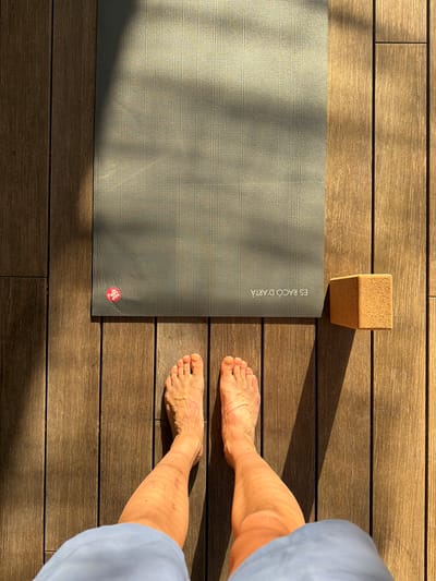 The image shows a top-down view of a yoga mat and a yoga block placed on a wooden deck. The scene is lit by sunlight, creating shadows across the mat and deck. Two bare feet are visible at the bottom of the frame, positioned on the wooden deck in front of the yoga mat. The yoga mat is dark gray and has a textured surface, with a small red circular logo near the bottom left corner and text that reads "ES RACO D'ARTÀ" printed in white. A light brown, rectangular yoga block is situated to the right of the yoga mat. The wooden deck is composed of parallel planks and appears to be outdoors. The presence of the text "ES RACO D'ARTÀ" on the yoga mat indicates that the scene is located at or related to "Es Racó d'Artà", which is a place in Artà, Spain.