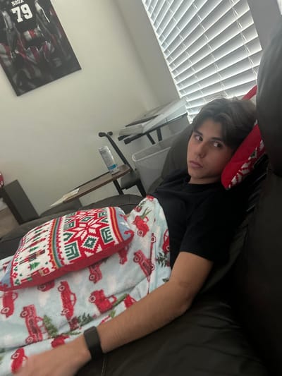 A person lies on a brown leather couch, covered in a light blue blanket with a red truck pattern. A pillow with a fair isle design is next to the person. The person wears a black shirt and a watch. In the background, there are closed blinds, a desk with a fan, and a poster with a sports figure. The scene is bathed in natural light from the window.