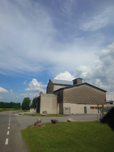 The image depicts an industrial building with a corrugated metal roof and white brick walls, located in Alavere, Estonia. A paved road with white dashed lines curves around the building. The building appears to be partially under construction or in disrepair, with exposed brickwork and some missing sections. Several large rocks are scattered on the grassy area in front of the building. The sky is partly cloudy with a mix of blue sky and white clouds. The surrounding environment includes green grass and trees in the distance. The overall scene suggests an industrial or rural setting.