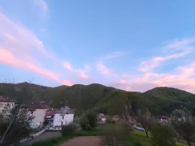 The image shows a landscape in Srednogortsi, Bulgaria, featuring a group of buildings situated at the foot of a range of green, tree-covered hills. The sky is blue with pink-tinged clouds. The buildings appear to be residential houses with varying roof colors, and some have chimneys. Trees and bushes surround the buildings. The foreground contains a patch of brown ground.