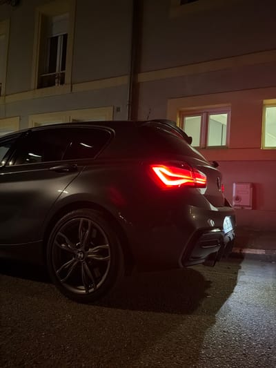 A gray BMW automobile is parked on a paved surface, likely a street or driveway, beside a building at night. The rear taillight of the car is illuminated. The building has windows with beige frames. A utility box is affixed to the wall of the building. The photograph appears to have been taken at ground level. Based on the image's provenance, this scene is potentially located in Villerupt, France.